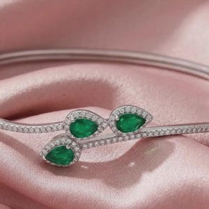 May include: A silver-toned hinged bangle bracelet featuring a bypass design adorned with three pear-shaped green gemstones. The band and the settings surrounding the green stones are encrusted with small, clear, sparkling pavé-set crystals.