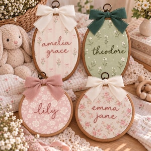 Könnte beinhalten: Vier hölzerne Stickrahmen mit Stoffeinsätzen mit Blumenmustern und gestickten Namen. Zwei sind oval mit weißen und grünen Schleifen, zwei sind rund mit rosa und weißen Schleifen und zeigen die Namen amelia grace, theodore, lily und emma jane.