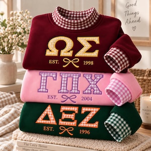 Könnte beinhalten: Ein Stapel aus drei langärmeligen Sweatshirts in Kastanienbraun, Rosa und Dunkelgrün. Jedes hat einen Stehkragen mit Vichy-Muster, passende Bündchen, gestickte griechische Buchstaben mit Vichy-Füllung, ein EST.-Datum und ein kleines Schleifensymbol.
