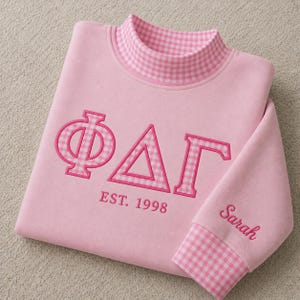 Puede incluir: Una sudadera rosa claro con cuello alto de patrón de cuadros vichy rosa y blanco. El frente muestra las letras griegas Phi Delta Gamma en aplique vichy con bordado rosa, y el texto EST. 1998. El nombre Sarah está bordado en la manga.