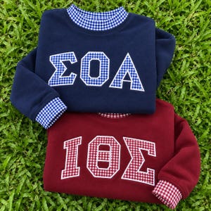 Könnte beinhalten: Ein marineblaues Sweatshirt mit blau-weiß kariertem Kragen, Bündchen und griechischen Buchstaben ΣΘΑ, über einem kastanienbraunen Sweatshirt mit rot-weiß kariertem Kragen, Bündchen und griechischen Buchstaben ΙΘΣ. Beide haben weiße Buchstabenränder.