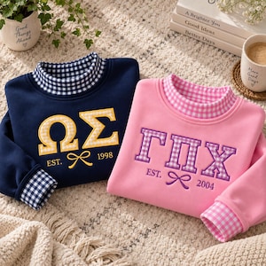 Puede incluir: Una sudadera azul marino con cuello y puños de cuadros vichy azules y blancos, con letras griegas amarillas y un lazo con el texto EST. 1998, junto a una sudadera rosa con cuello y puños de cuadros vichy rosas y blancos, con letras griegas moradas y un lazo con el texto EST. 2004.