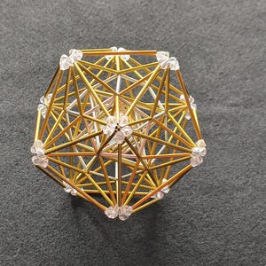 Könnte beinhalten: Eine geometrische Skulptur aus dünnen goldenen und silbernen Metallstäben, die in einem komplexen polyedrischen Gitter angeordnet sind. Die Eckpunkte der Struktur sind an jedem Schnittpunkt mit kleinen, klaren, facettierten Kristallperlen verbunden.