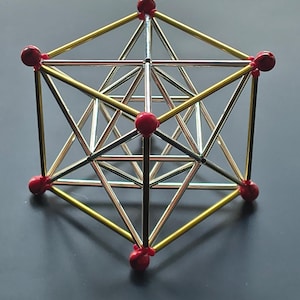 Könnte beinhalten: Ein geometrisches Modell aus Metallstäben und roten kugelförmigen Verbindungsstücken. Die Struktur besteht aus einem großen äußeren Würfelrahmen mit goldfarbenen Stäben und einem inneren Gerüst aus silberfarbenen Stäben in komplexer Form.