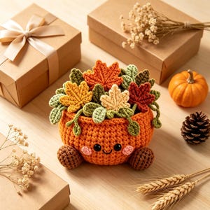 Puede incluir: Una maceta de ganchillo con forma de calabaza naranja, con una cara sonriente, mejillas rosadas y dos pies marrones. La maceta está llena de varias hojas de otoño de ganchillo en tonos naranja, amarillo, crema y verde, junto a enredaderas.