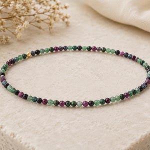May include: A delicate beaded bracelet featuring small, faceted, round gemstones in alternating shades of deep green, dark blue, and magenta. The strand includes a single, small, polished gold-toned metal bead positioned near the top left section.