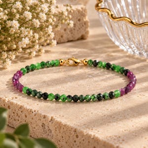 Pode incluir: Uma pulseira delicada de contas com pequenas esferas facetadas em tons alternados de verde vibrante, preto profundo e magenta rico. A pulseira é fixada com um fecho lagosta de metal em tom dourado polido, criando um acessório refinado.