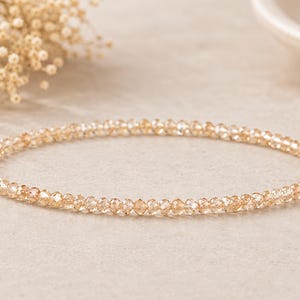 Könnte beinhalten: Ein zartes, kreisförmiges Armband, das aus zahlreichen kleinen, facettierten champagnerfarbenen Kristallen besteht. Die Perlen sind in einer durchgehenden, gleichmäßigen Linie aufgereiht und bilden eine schimmernde Oberfläche.