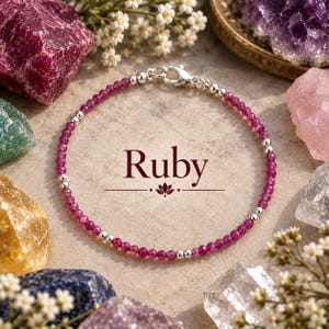 Op de afbeelding: Een delicate armband met kleine, gefacetteerde, donkerroze robijnkralen afgewisseld met gepolijste zilverkleurige metalen bolletjes. De armband is voorzien van een zilverkleurige karabijnsluiting. De tekst Ruby staat boven een klein lotusicoon.
