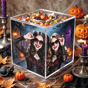 May include: A clear acrylic cube container filled with assorted Halloween candies, featuring a printed image of two individuals wearing black lace tops and sunglasses. The cube is surrounded by small orange pumpkins, a black cat figurine, and purple candles.