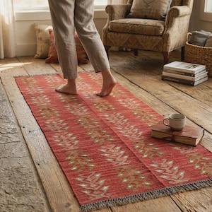 May include: A rectangular runner rug featuring a vibrant red base with repeating vertical patterns in muted green and cream tones. The rug has a textured weave and short fringe detailing along the edges, providing a flat surface for bare feet.