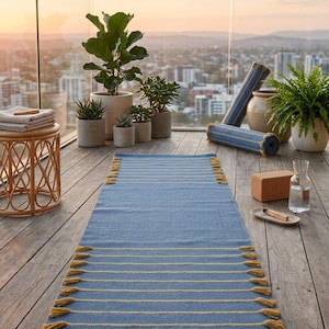 May include: A rectangular blue yoga mat featuring horizontal yellow stripes and decorative yellow tassels along the short edges. The mat is laid flat on a wooden surface, with a brown leather-like brand tag attached to one corner of the mat.
