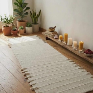 May include: A rectangular off-white woven runner rug featuring a textured surface with horizontal ribbed stripes and decorative fringe tassels along the short edges. The rug is crafted from a light-colored fabric with a consistent weave pattern.