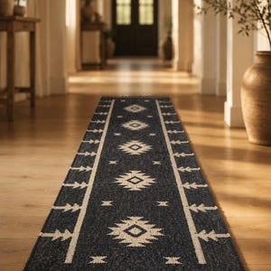 May include: A long, rectangular runner rug featuring a dark charcoal gray base with a repeating cream-colored geometric diamond pattern. The rug is bordered by a cream-colored line with tree-like motifs and finished with short, gray fringe at the ends.