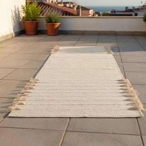 May include: A rectangular off-white runner rug featuring a woven texture and decorative fringe detailing along both short ends. The rug is positioned lengthwise on a tiled surface, displaying a consistent light cream color throughout its surface.