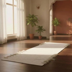 May include: A rectangular off-white woven runner rug featuring a textured surface and decorative fringe along the short edges. The rug is positioned on a dark wooden floor in a room with natural light streaming through large windows.