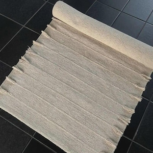 May include: A rectangular off-white woven rug featuring a textured surface with vertical ribbed stripes and short fringe detailing along both side edges. The rug is partially rolled at the top end, displaying a consistent knit pattern throughout.