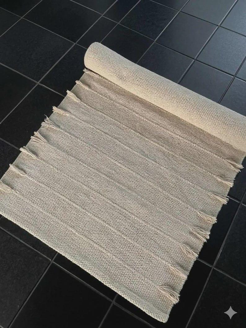 May include: A rectangular off-white woven rug featuring a textured surface with vertical ribbed stripes and short fringe detailing along both side edges. The rug is partially rolled at the top end, displaying a consistent knit pattern throughout.