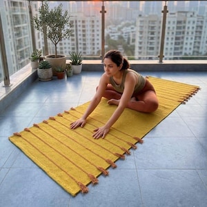 May include: A rectangular mustard yellow yoga mat featuring a textured surface with evenly spaced horizontal lines and decorative tassels along both long edges. The mat is laid flat on a tiled floor, providing a surface for exercise and movement.