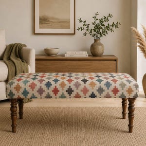 May include: A rectangular upholstered bench featuring a repeating geometric cross pattern in shades of red, blue, yellow, and purple on a cream background. The bench is supported by four dark brown turned wooden legs with a classic spindle design.