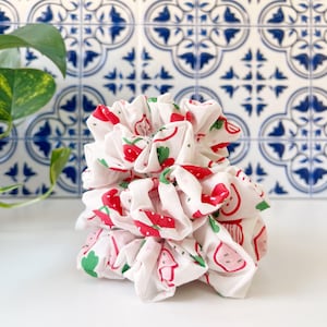 Peut inclure: Plusieurs chouchous blancs ornés d'un motif de fraises rouges et vertes. Le tissu est froncé et superposé, créant un effet volumineux. Le fond présente un motif de carreaux bleus et blancs.