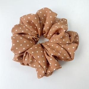 Peut inclure: Grosse chouchou marron avec un motif à pois blancs. Le tissu est froncé et plié, créant une forme circulaire volumineuse. Le chouchou est conçu pour tenir les cheveux et convient à diverses coiffures.