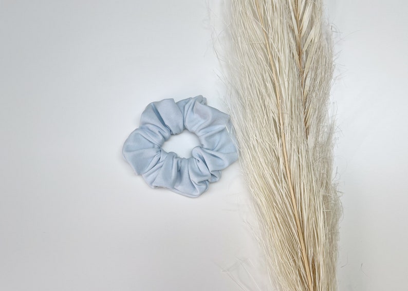 May include: A light blue scrunchie made of soft fabric. The scrunchie is in the shape of a circle and has a gathered look.