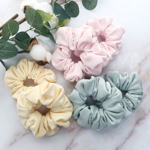 May include: Four scrunchies in different colours: light pink, light yellow, light green, and a darker shade of green. The scrunchies are made of soft fabric and are perfect for holding hair in place.