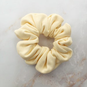 May include: A pale yellow scrunchie made of soft fabric. The scrunchie is gathered in a circle and has a smooth, even texture.
