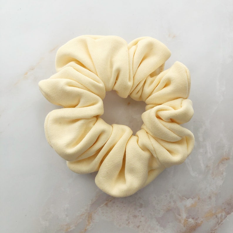 May include: A pale yellow scrunchie made of soft fabric. The scrunchie is gathered in a circle and has a smooth, even texture.