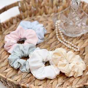 May include: Five fabric scrunchies in a wicker basket. The scrunchies are in pastel colours: light blue, pink, light green, white, and cream.