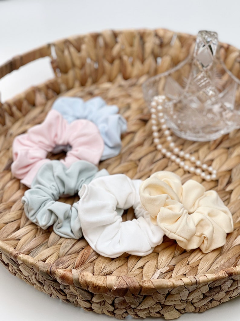 May include: Five fabric scrunchies in a wicker basket. The scrunchies are in pastel colours: light blue, pink, light green, white, and cream.