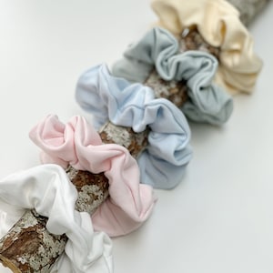 May include: A set of six scrunchies in various pastel colours, including white, light blue, pink, and cream. The scrunchies are arranged on a piece of driftwood.