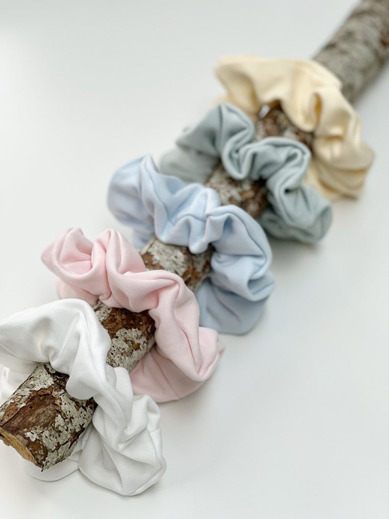 May include: A set of six scrunchies in various pastel colours, including white, light blue, pink, and cream. The scrunchies are arranged on a piece of driftwood.