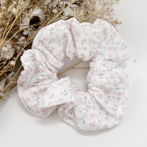May include: White scrunchie featuring a dainty floral design in pink and grey. The fabric is gathered and elasticated, forming a voluminous, circular shape. This hair accessory is ideal for adding a touch of style to any hairstyle.