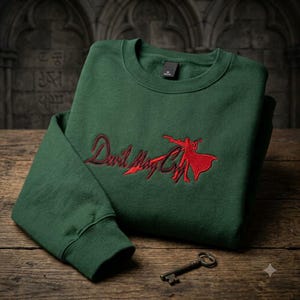 Peut inclure: Un sweat-shirt à col rond vert forêt avec une broderie rouge sur la poitrine. La broderie représente le texte Devil May Cry accompagné de la silhouette d'un personnage tenant une arme. Une petite clé en métal est placée sous le vêtement.