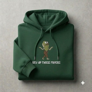 Può includere: Una felpa con cappuccio verde scuro caratterizzata da una grafica ricamata di un personaggio pesce dei cartoni animati che indossa pantaloni marroni. Sotto il personaggio, il testo "REV UP THOSE FRYERS" è scritto in lettere maiuscole.