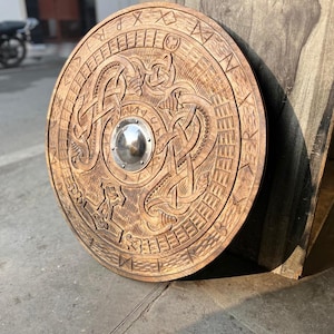 May include: A circular wooden shield featuring intricate carved knotwork patterns and a central metallic boss. The outer rim is decorated with engraved runic symbols, and the wood displays a natural brown grain with dark stained details.
