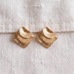 May include: A pair of gold-toned stud earrings featuring a square shape with a concave center and a textured, ribbed surface. The earrings have a metallic finish and a geometric design, showcasing a repeating pattern of fine, parallel lines.