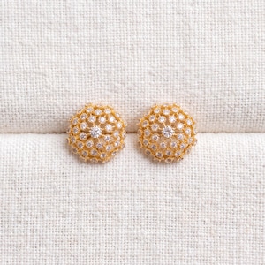 May include: A pair of gold-tone stud earrings featuring a domed, circular design. Each earring is densely encrusted with numerous small, clear, round-cut stones arranged in a radial pattern, creating a textured, sparkling surface.