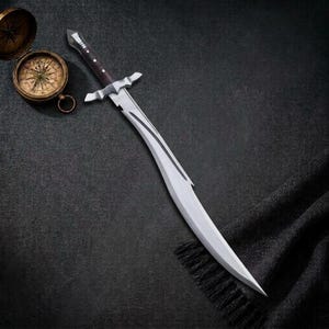 May include: A curved steel sword with a dark brown handle and a silver crossguard, positioned next to an open antique brass compass with a detailed face featuring black markings and a needle on a dark textured surface.