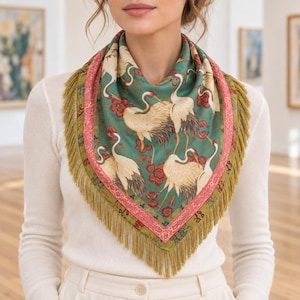 May include: A square scarf featuring a teal green background with a pattern of white cranes and red floral accents. The scarf is bordered by a pink patterned trim and finished with long, olive green fringe along the edges of the fabric.