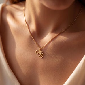 Peut inclure: Un collier doré composé d'une fine chaîne à maillons carrés et d'un pendentif formé par les lettres cursives M et E entrelacées. Le pendentif est suspendu au centre de la chaîne, qui repose au niveau de la clavicule dans un style épuré.