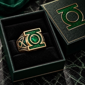 May include: A gold-toned ring featuring a central circular green gemstone and the Green Lantern symbol, set within a dark green textured box. The ring band displays intricate geometric cutouts and green inlay details, matching the box lid design.