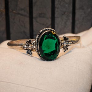 May include: A silver-toned cuff bracelet featuring a large, oval-shaped, faceted emerald green stone at the centre. The stone is framed by a twisted rope border, and the band is accented on both sides by small, detailed metal bee motifs.