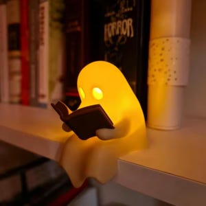 Op de afbeelding: Een gloeiende, gele, spookvormige lamp die op de rand van een plank zit. Het spookfiguur heeft twee ronde oogopeningen en houdt een klein, donkerkleurig open boek in de handen, verlicht door het warme licht dat van binnenuit het lichaam komt.