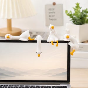 Op de afbeelding: Vier kleine witte ganzenbeeldjes met oranje snavels en poten zitten op de bovenrand van een laptopscherm. Eén gans ligt plat, één hangt ondersteboven, één zit rechtop met een hand aan de kin en één klampt zich vast aan de zijkant van het scherm.