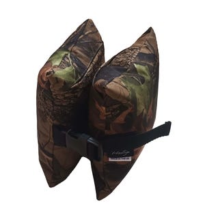 May include: A dual-chamber shooting rest bag featuring a brown, green, and black camouflage pattern. The two bags are connected by a black nylon strap with a plastic side-release buckle. A small white label reads "Hastags" and "MADE IN THE UK".