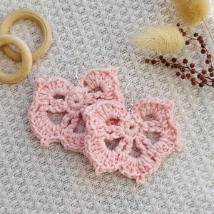 May include: A pair of light pink crochet earrings featuring a floral-inspired openwork pattern. Each earring is crafted from yarn and attached to a silver-toned metal hook. The design consists of a central circular element with five radiating petals.