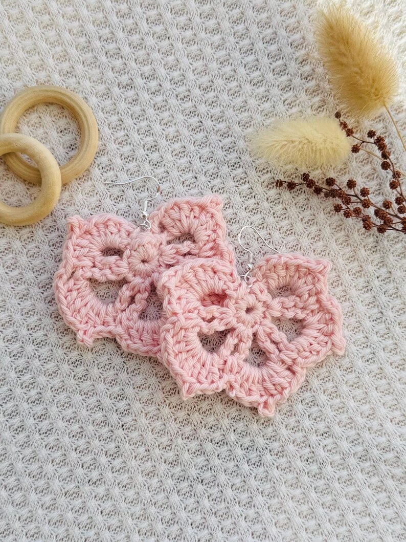 May include: A pair of light pink crochet earrings featuring a floral-inspired openwork pattern. Each earring is crafted from yarn and attached to a silver-toned metal hook. The design consists of a central circular element with five radiating petals.